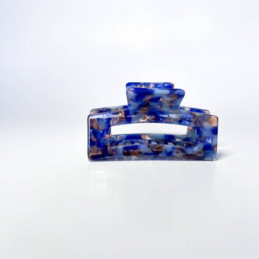 A large, translucent light cobalt blue claw clip is shown against a plain white background. The clip has a rectangular shape with rounded edges and is slightly open, revealing its spring mechanism and gripping teeth.